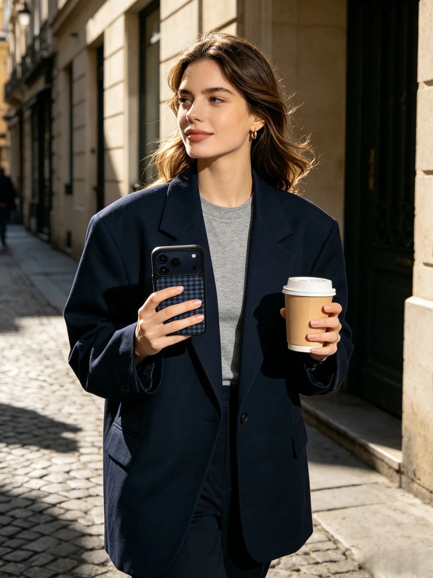 An edgy streetstyle photo of a woman wearing an oversized navy blue blazer walking down a cobblestone European street, holding a coffee cup and a blue plaid dachshund phone case with a black bumper.