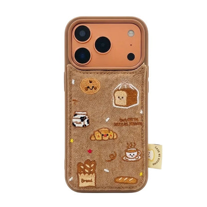 A tan denim iPhone case featuring detailed 3D embroidered patches of croissants, a milk carton, coffee, and baguettes.