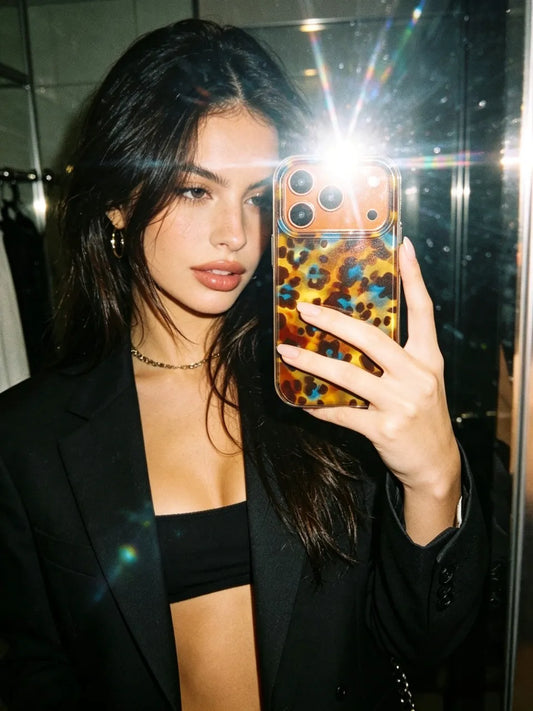 A young woman taking a mirror selfie with a flash, showcasing a trendy CasePoppi leopard print phone case paired with a black blazer.