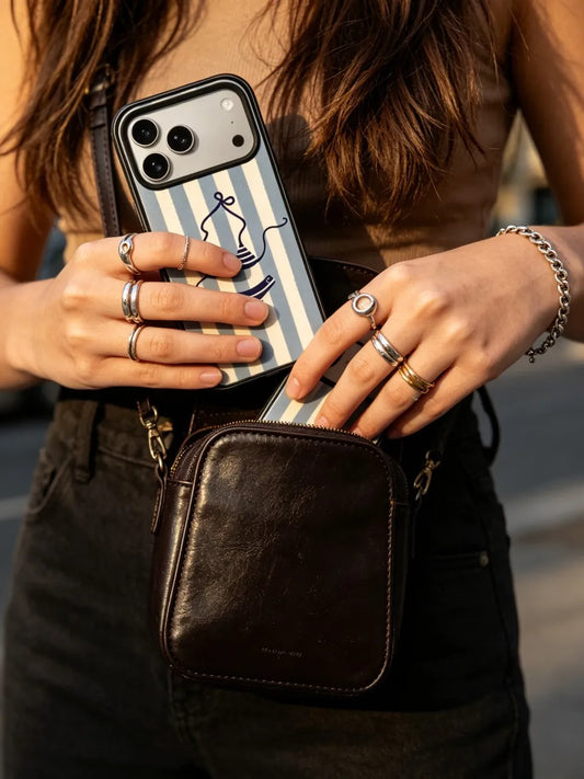 Detail shot of a person putting a blue and white striped CasePoppi phone case into a black leather crossbody bag, streetstyle fashion look.