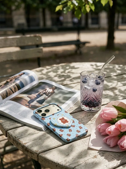 A CasePoppi blueberry teddy bear gingham phone case placed on a rustic wooden outdoor table with a sparkling blueberry drink and pink tulips.
