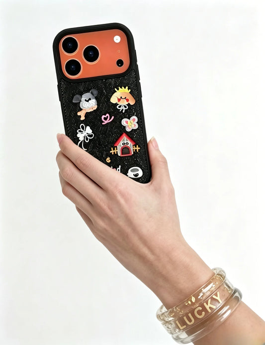 A hand wearing a clear glitter 'LUCKY' bangle holds a CasePoppi black textured phone case adorned with 3D cute cartoon puppies, a dog house, and ribbon bow charms.
