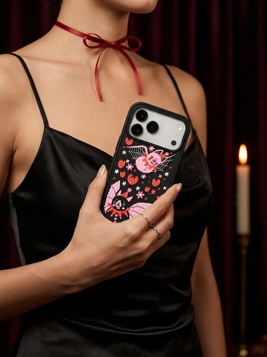 A close-up of a woman in a black silk slip dress and red ribbon choker holding the CasePoppi spooky cute gothic Strawberry Bat Duo phone case.