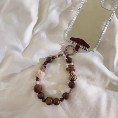 CasePoppi fluffy pompom and wood beaded phone strap attached to a ripple-textured ripple phone case, resting on soft white bedding, cozy aesthetic.