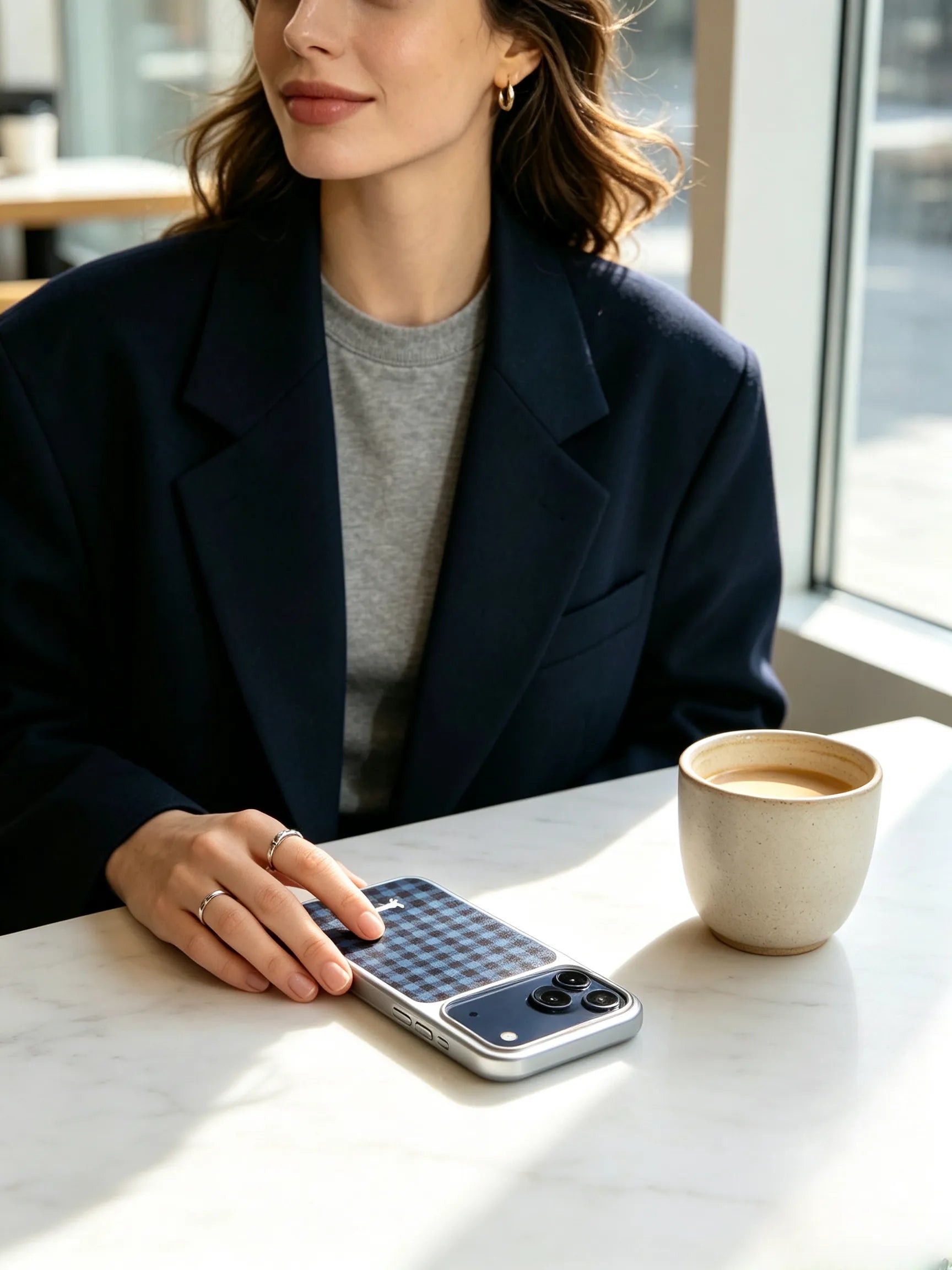 A stylish cafe aesthetic photo of a woman in a navy blazer sitting at a marble table, resting her phone with a blue plaid dachshund pattern and silver camera bumper next to a cup of coffee.