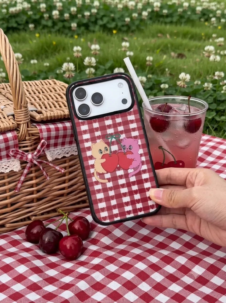 A still life aesthetic photo of a red and white plaid gingham phone case with a cute cherry character design, set on a matching picnic blanket with fresh cherries and an iced drink.