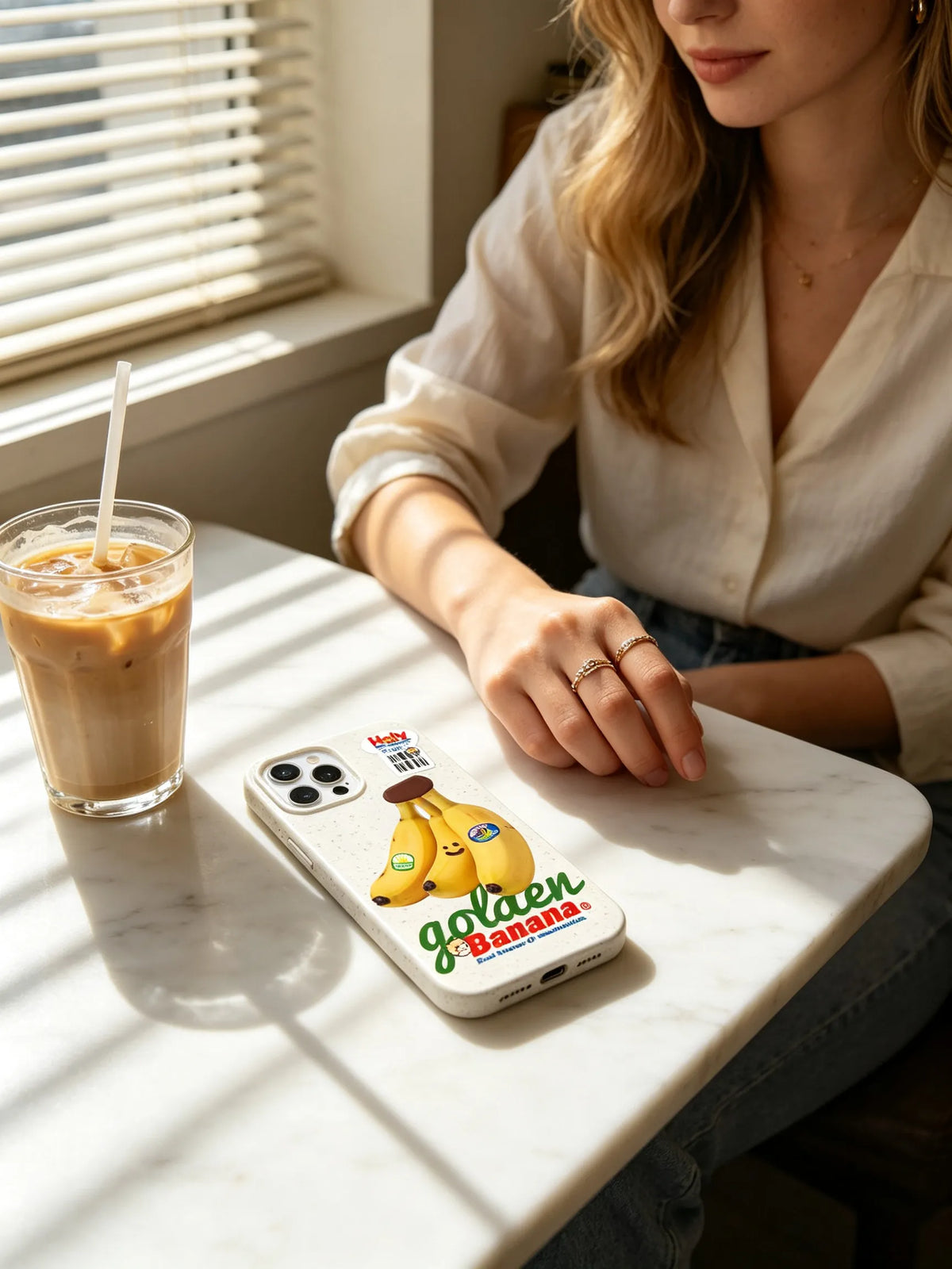 A sunlit aesthetic table top setup showing an off-white phone case with a retro golden banana illustration lying next to a glass of iced coffee near window blinds.