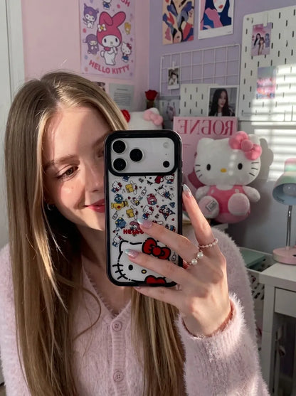 A cozy dorm room mirror selfie of a girl with long hair wearing a pastel pink cardigan, showcasing her colorful Hello Kitty phone case with plushies in the background.