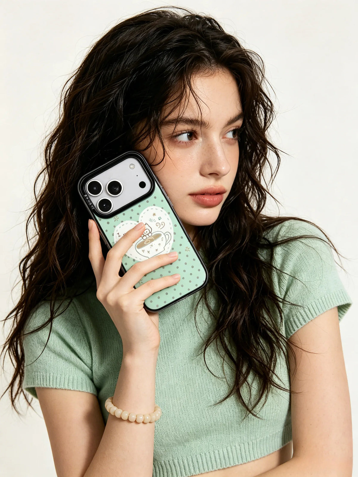 A close-up lifestyle photo of a young woman with wavy long dark hair, holding a mint green and white checkered phone case in a cozy minimalist indoor setting.
