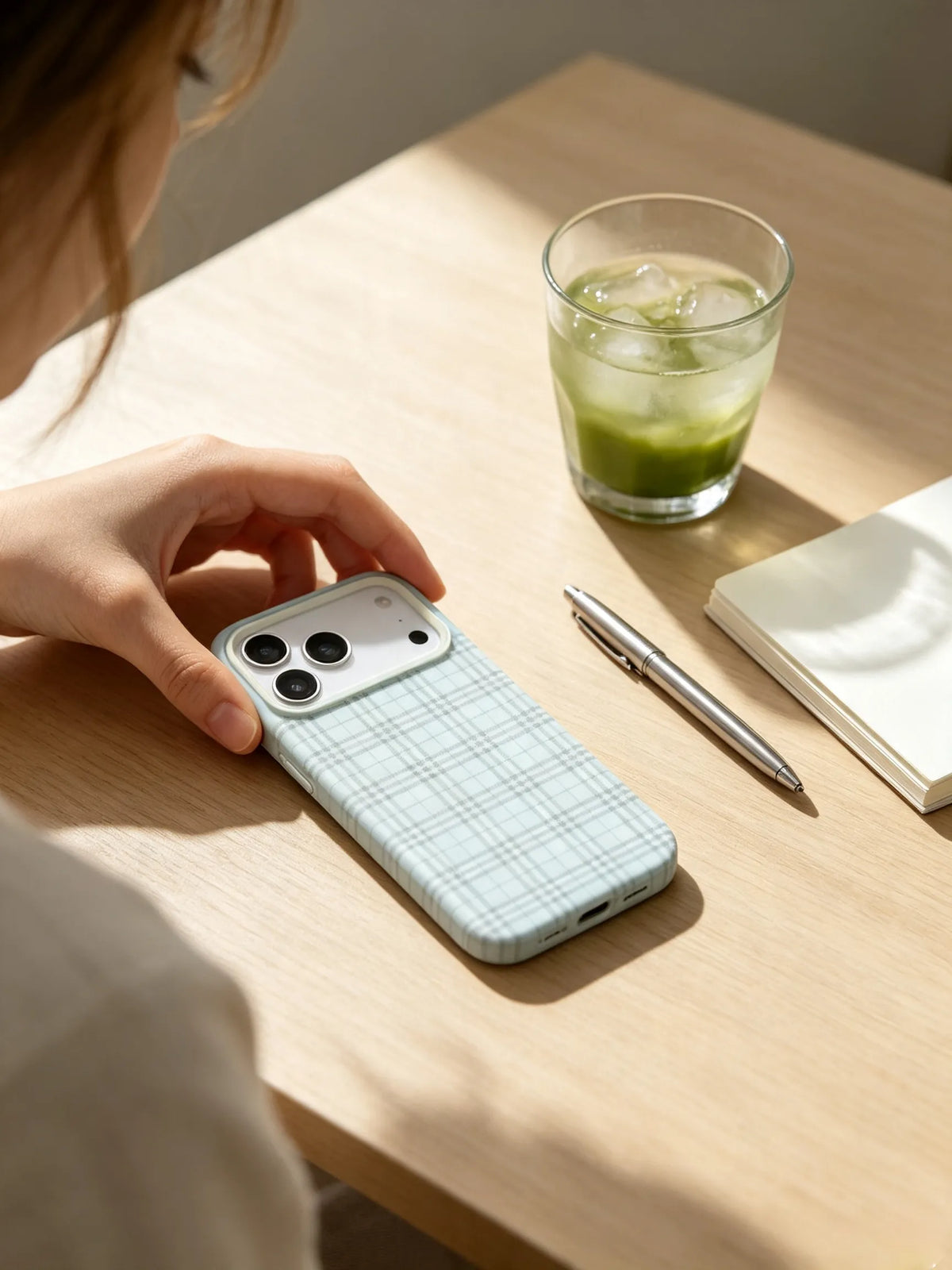 An aesthetic aesthetic desk setup photo of a light blue plaid phone case lying next to a glass of iced matcha latte and a notebook under warm sunlight.