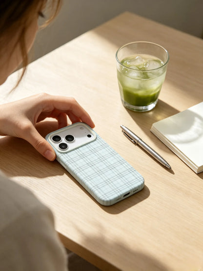 An aesthetic aesthetic desk setup photo of a light blue plaid phone case lying next to a glass of iced matcha latte and a notebook under warm sunlight.