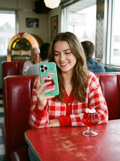 A retro 50s diner lifestyle photo of a woman in a red red-and-white checkered cardigan holding a mint green jelly pudding phone case next to a glass of red cherry jello dessert.