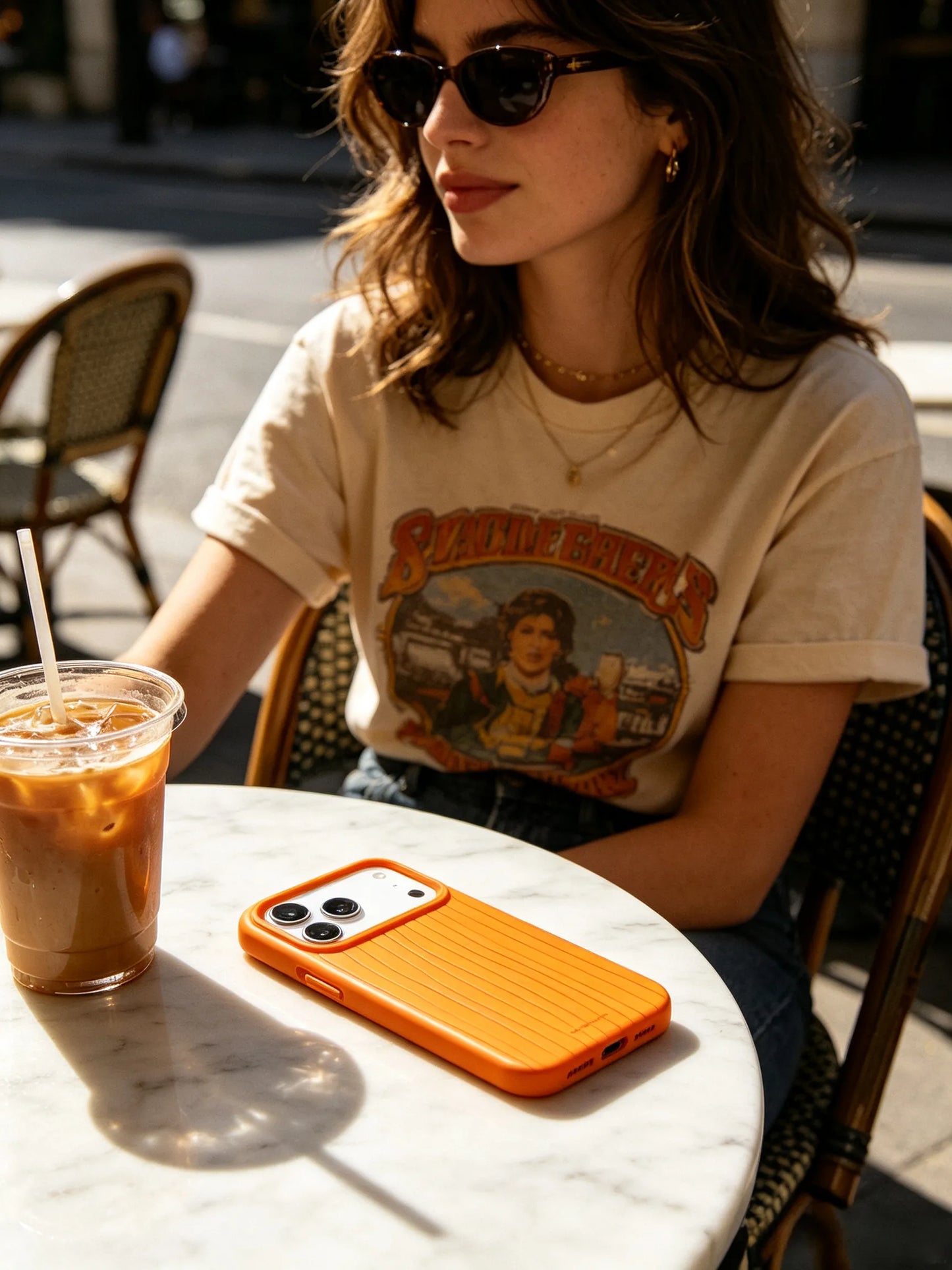 A sunny streetstyle photo of a woman in a graphic t-shirt sitting at an outdoor bistro table with an iced coffee, displaying a bright orange textured phone case.